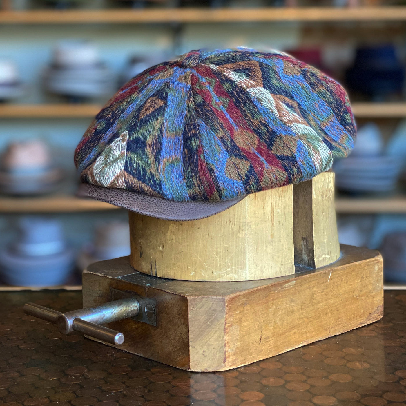 90s Sweater Hatteras features a multi-colored textured fabric with a soft pattern and a contrasting solid brim, displayed on a wooden hat block in a cozy hat shop setting.