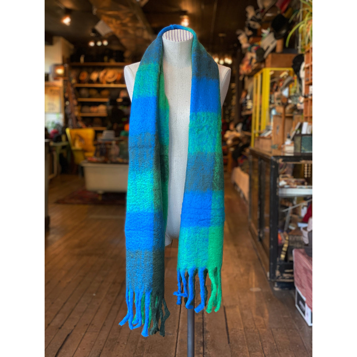 Colorblock Check Scarf in vibrant blue and green tones displayed on a mannequin in a cozy, rustic shop setting with wooden floors and shelves filled with accessories.