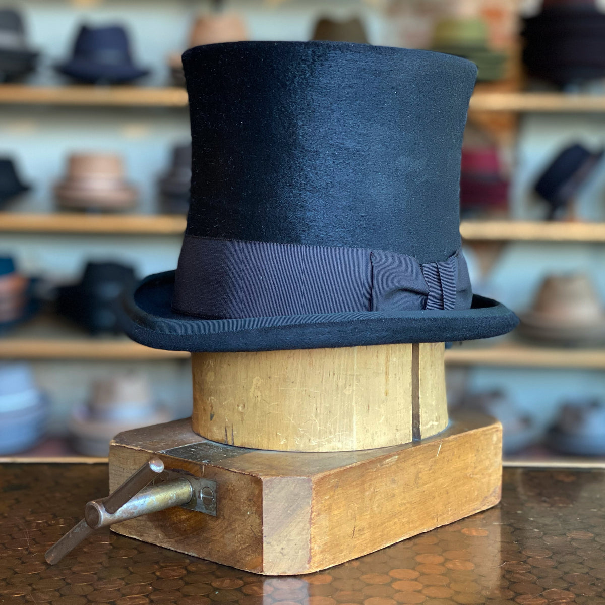 Classic black Paris Top Hat with a wide, dark ribbon band, displayed on a wooden hat block in a shop setting featuring various hats in the background.