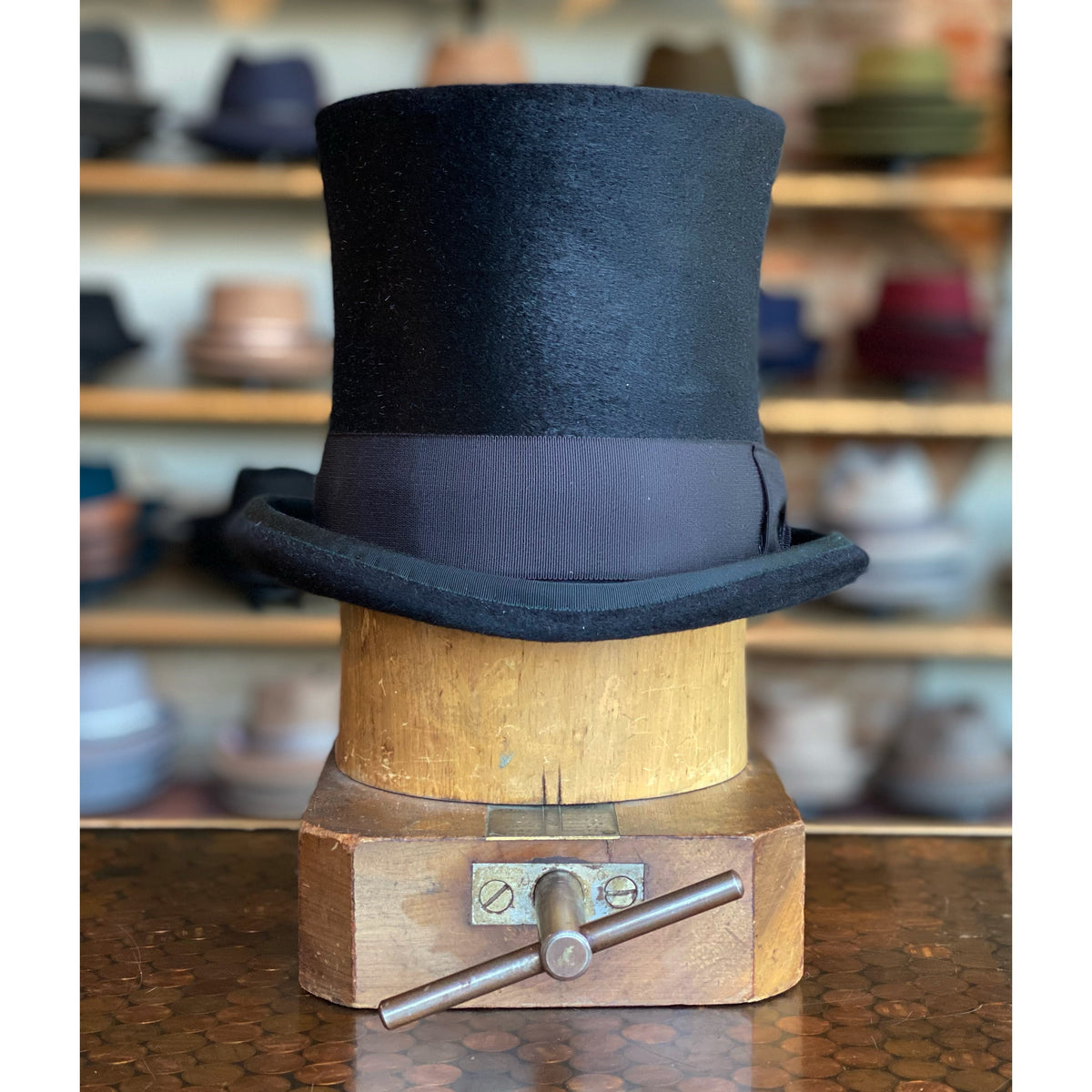 Black Paris Top Hat with wide brown band displayed on a wooden hat block, set against blurred shelves filled with various hats and caps in the background.