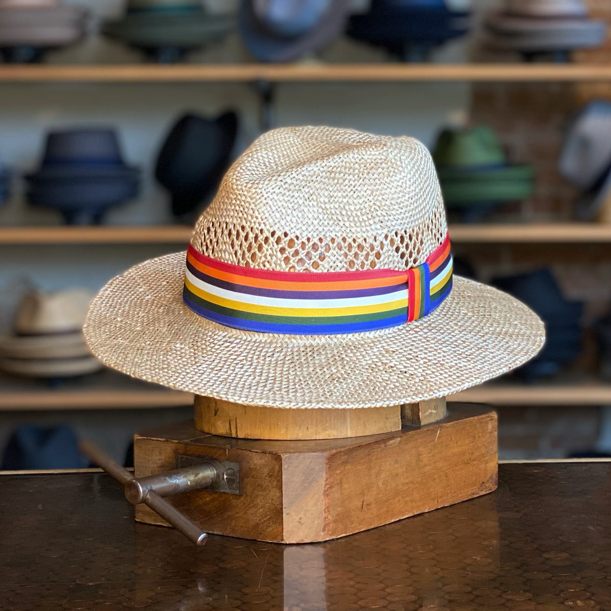 Pride Fedora straw hat featuring a colorful rainbow band, displayed on a wooden hat block in a shop with various hats in the background.