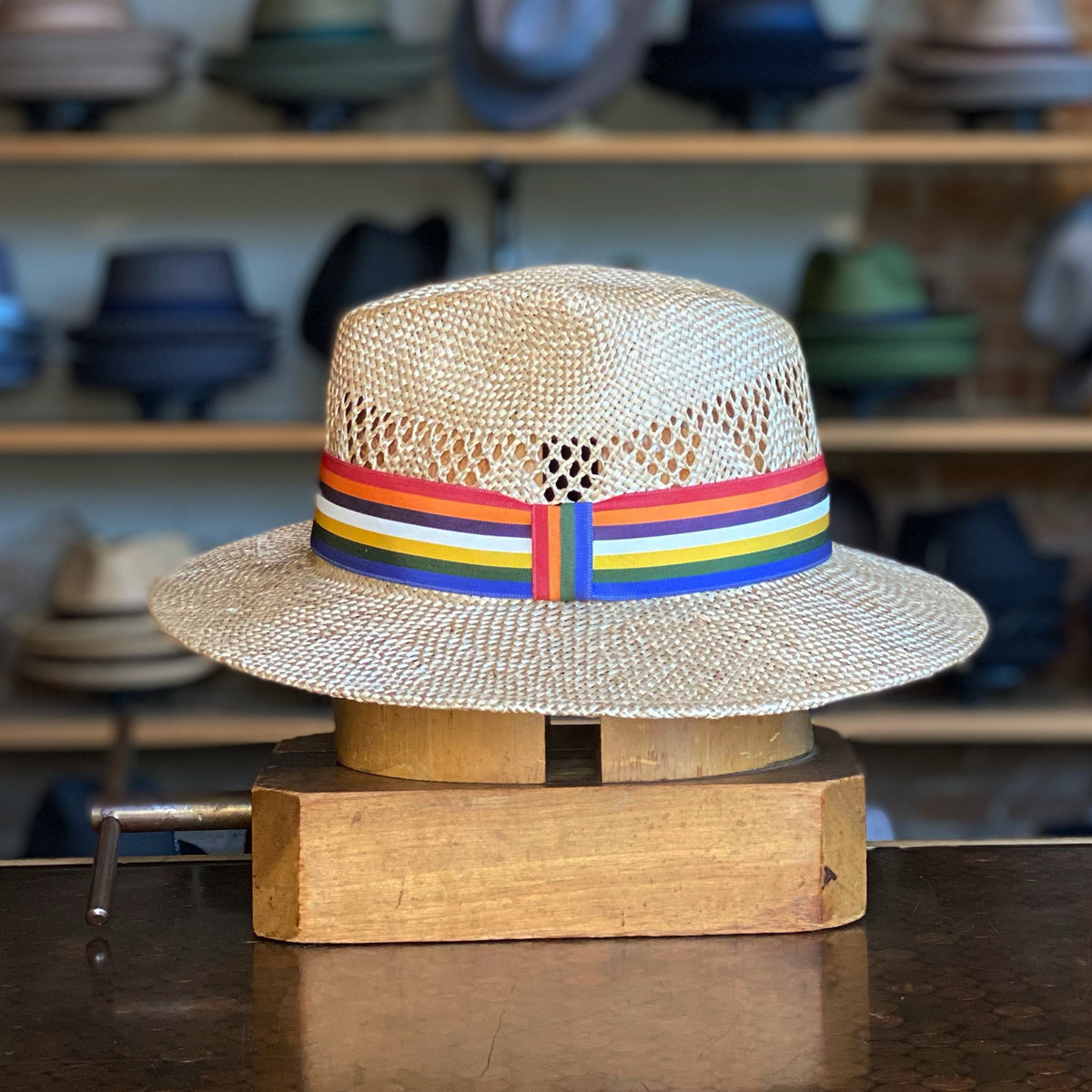 Pride Fedora featuring a woven straw design with ventilation cutouts and a colorful rainbow-striped band, displayed on a wooden hat stand in a hat shop setting.