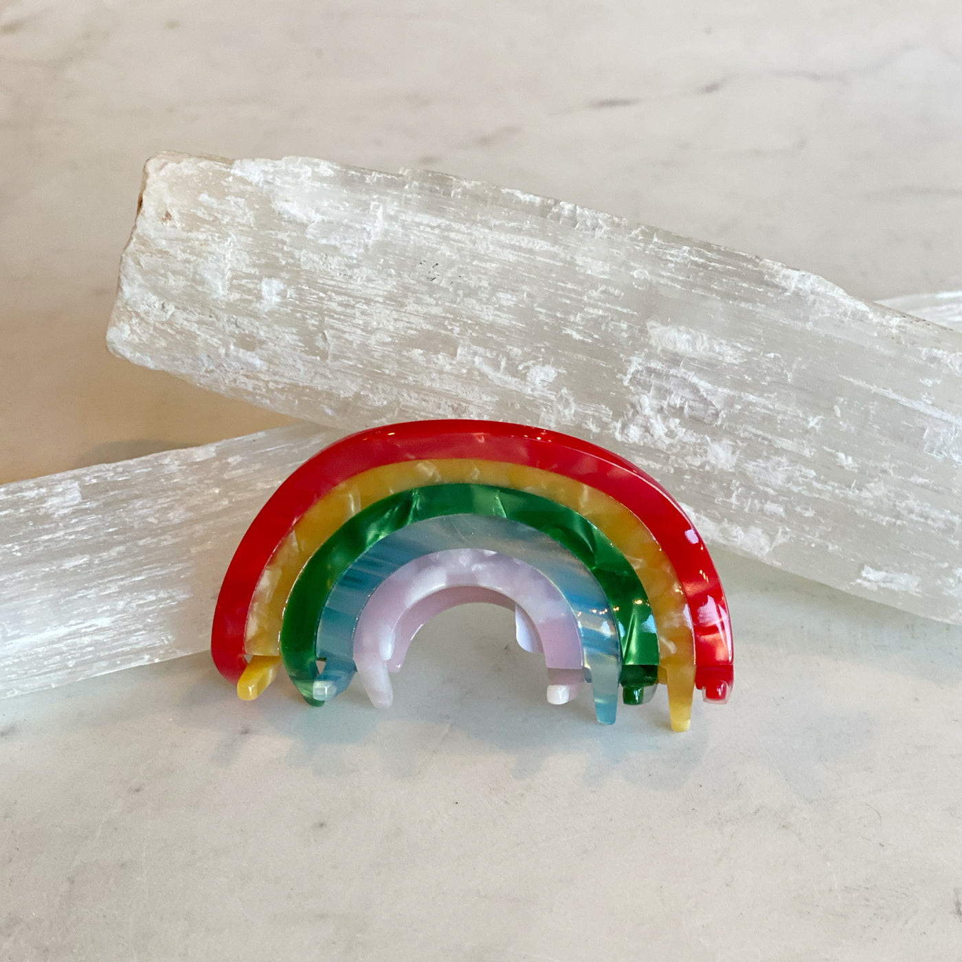 Rainbow Hair Clip featuring vibrant arches in red, yellow, green, and lavender colors, resting on textured white mineral crystals on a neutral surface.