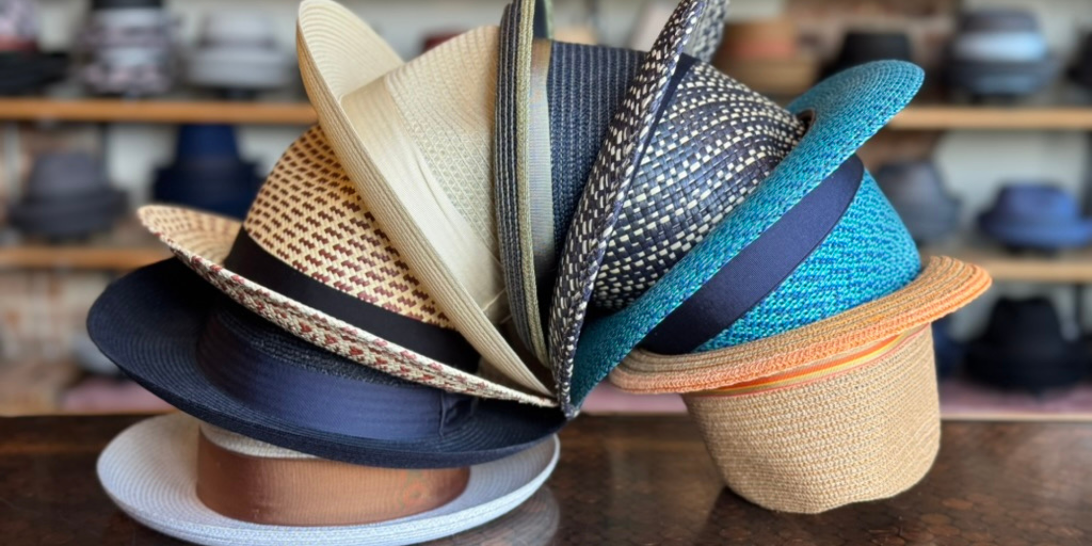 A colorful arrangement of various fedoras including summer braids splayed in a semi-circle on a wooden surface with blurred shelves of hats in the background.