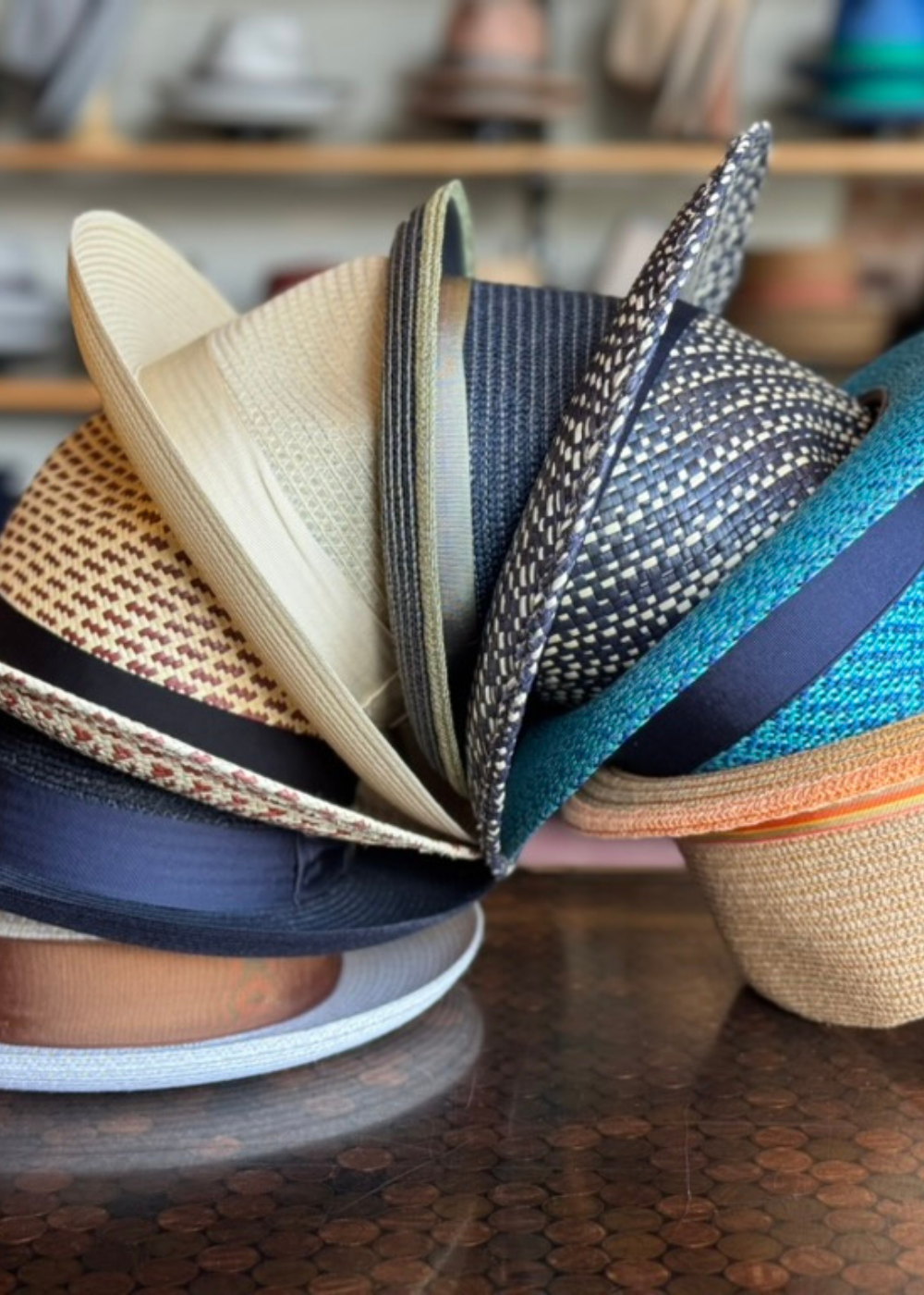 A close-up view of a variety of summer braid fedoras stacked featuring different colors and patterns against a blurred background of other hats on shelves.
