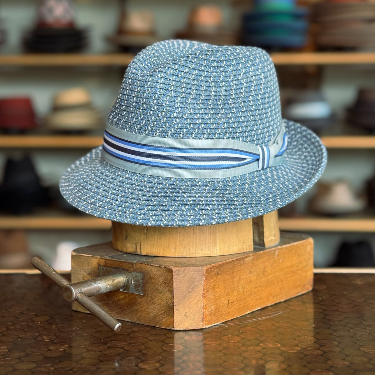 Salem blue woven fedora with a striped band, displayed on a wooden hat block in a shop with various hats in the background.