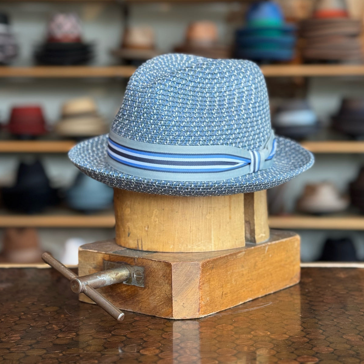 Salem blue woven fedora hat with striped ribbon band displayed on a wooden hat block in a hat shop with shelves of various hats in the background