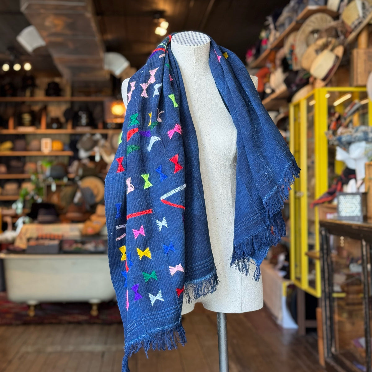 Vintage Indigo Mud Cloth Scarf No. 1 draped on a mannequin in a cozy, rustic shop setting, featuring deep indigo fabric with vibrant, colorful embroidered accents and fringed edges.