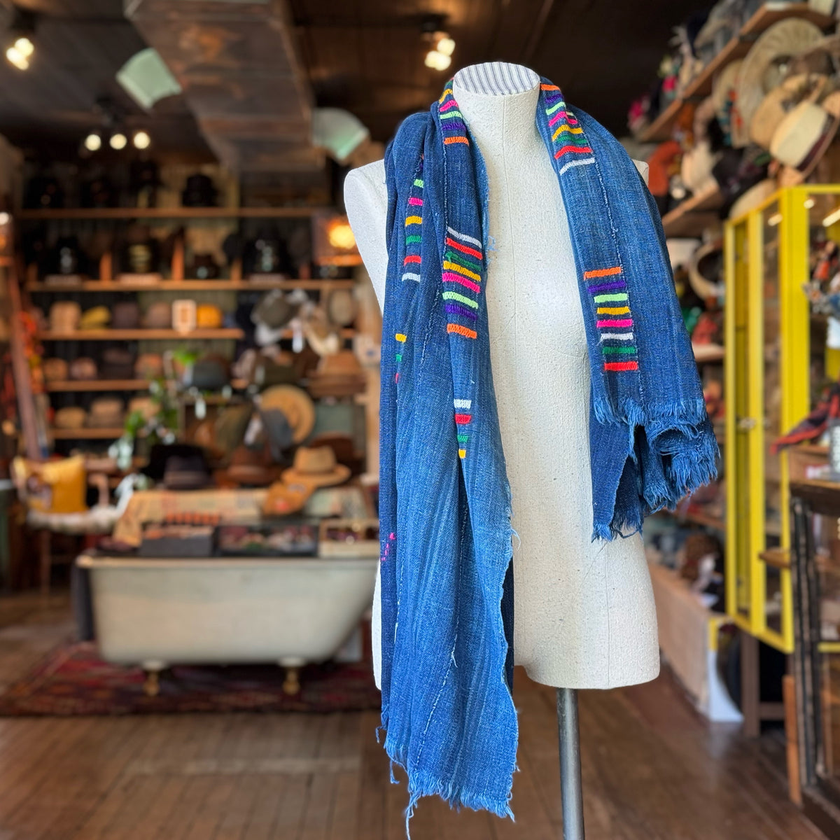 Vintage Indigo Mud Cloth Scarf No. 2 draped on a white mannequin in a rustic boutique, featuring deep blue fabric with vibrant multicolor accents and frayed edges.