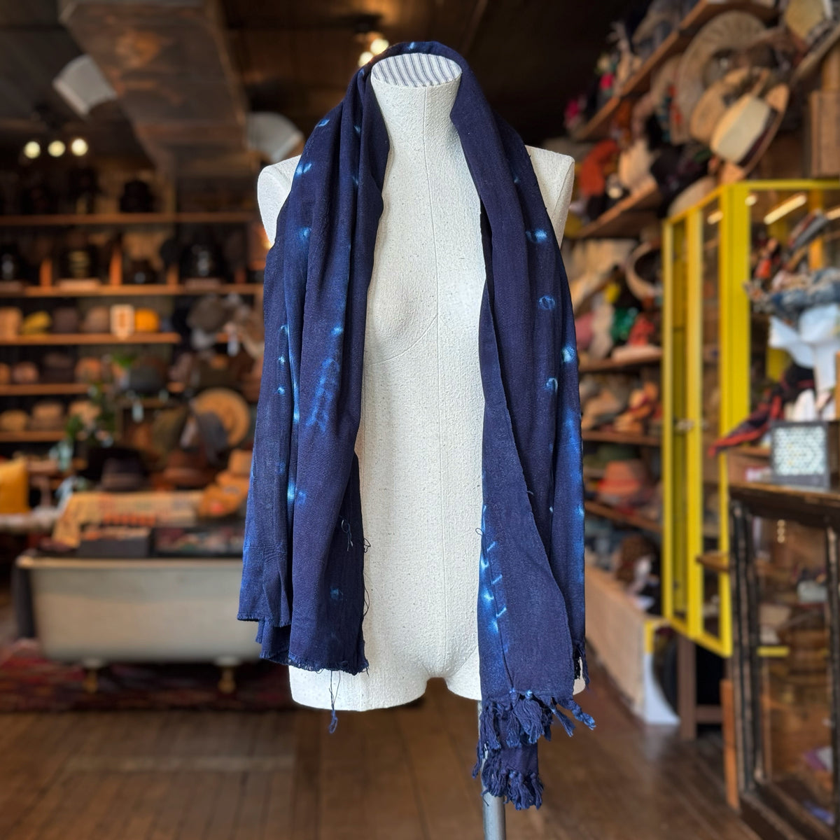 Vintage Indigo Mud Cloth Scarf No.6 draped on a mannequin in a rustic boutique setting, showcasing its deep indigo color and subtle mud cloth patterns with fringe detailing.
