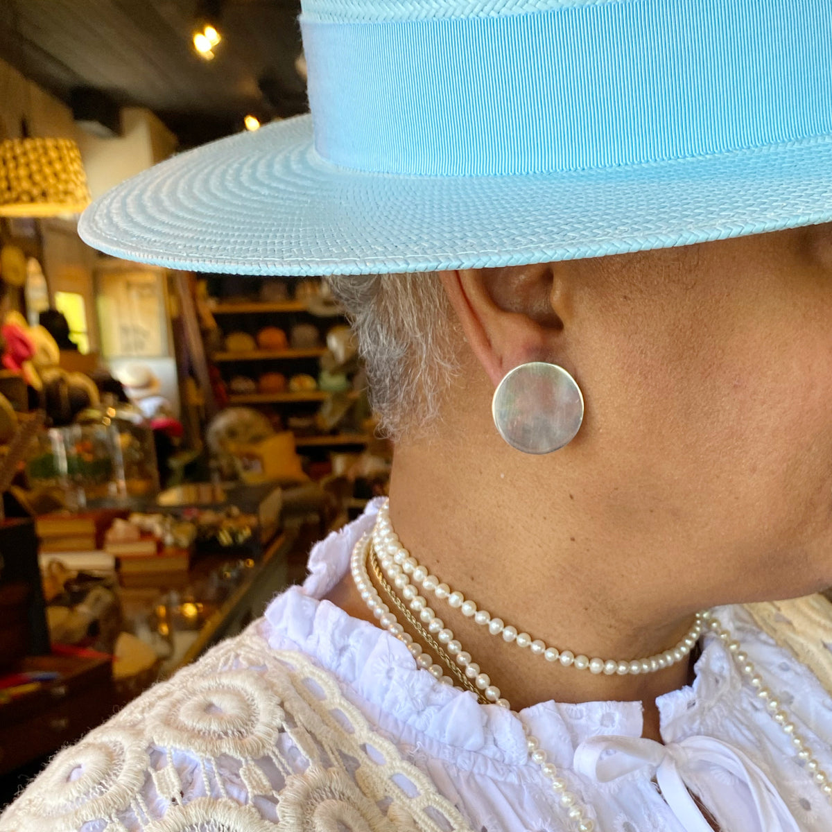 Close-up of a Jane wearing a light blue hat and a Sterling Disc Earring, paired with layered pearl necklaces and a textured white blouse in a cozy shop setting.