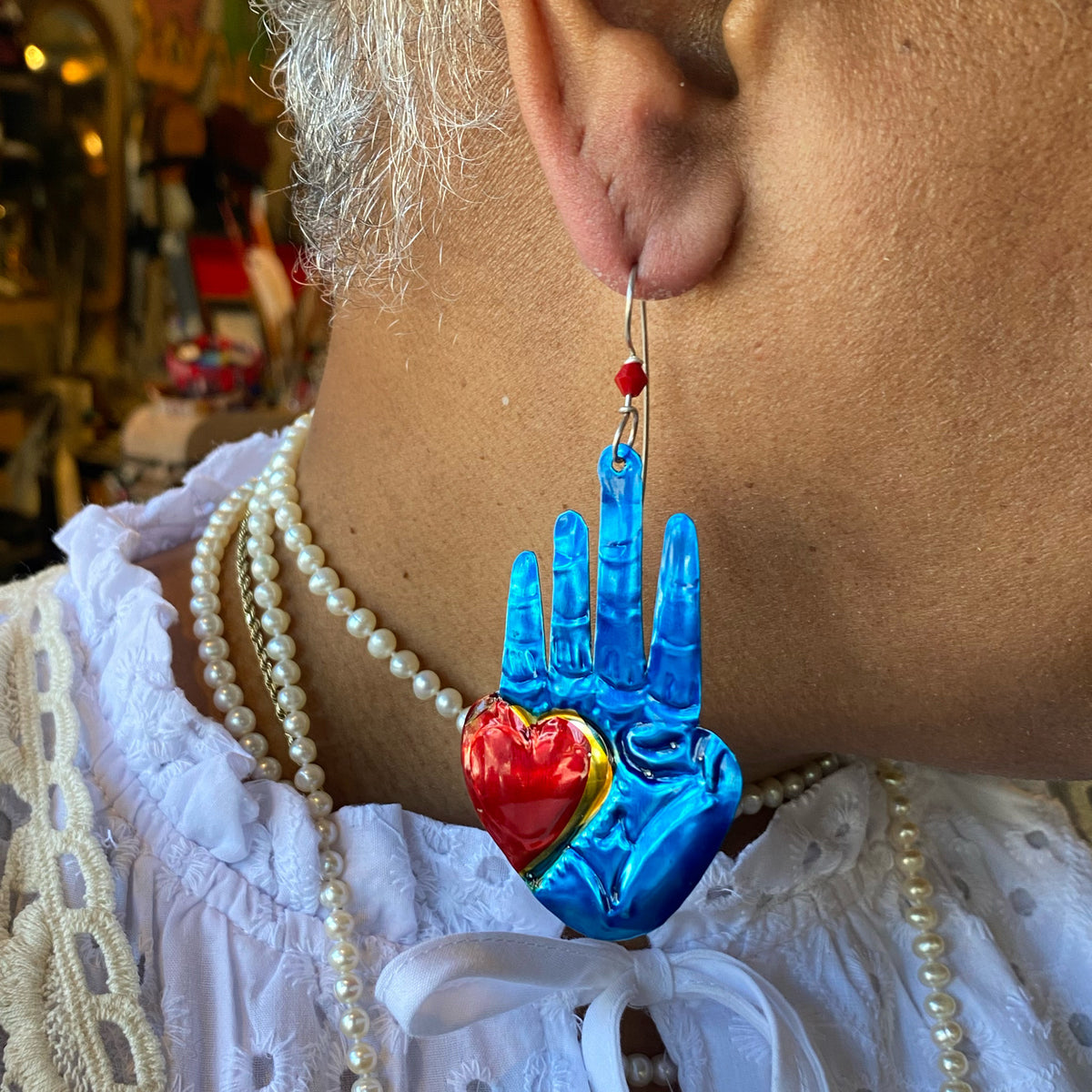 Close-up of Jane wearing a Blue Hand Earring designed as a shiny blue hand with a red heart detail, paired with a white lace top and pearl necklaces.