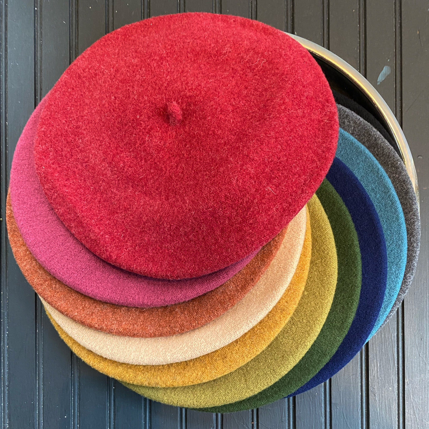 A colorful stack of Flora Beret hats in various shades including red, pink, orange, yellow, green, blue, and gray, displayed on a dark wooden surface.