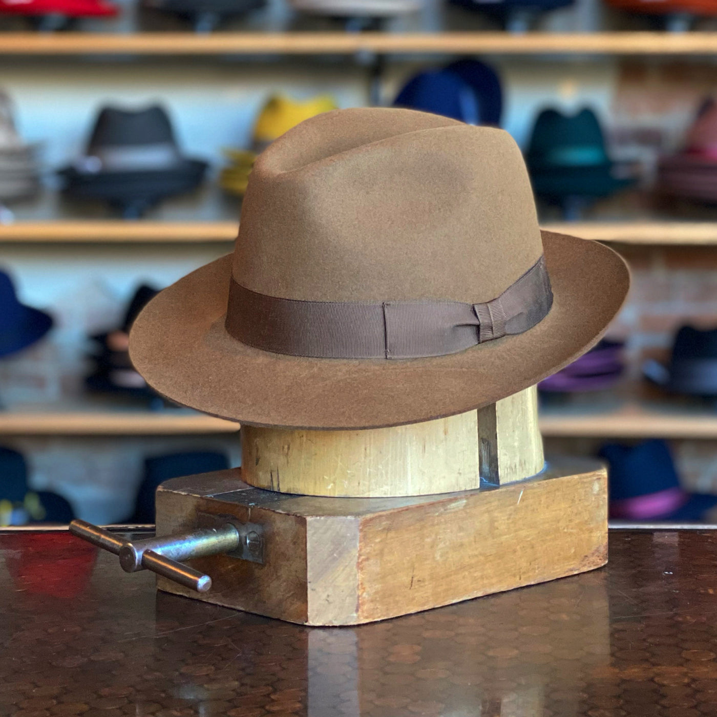 Garbo brown felt fedora with a dark brown ribbon band displayed on a wooden hat block in a hat shop with various hats in the background.