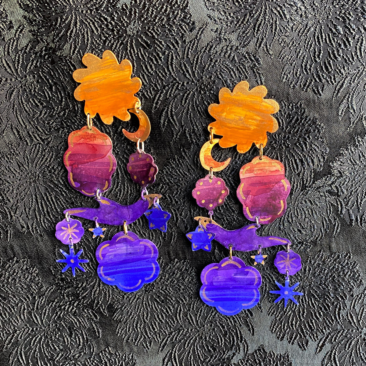 Pair of colorful Gilded Sunset Earring featuring orange, pink, and purple tones with celestial shapes including moons and stars on a textured black background.