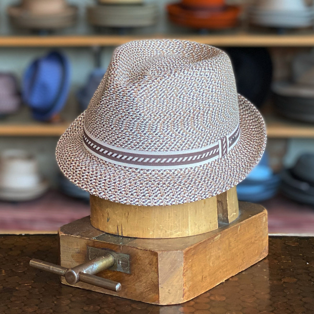 Mannes woven light brown fedora hat with a striped fabric band, displayed on a wooden hat block in a hat shop with shelves of various hats in the background.