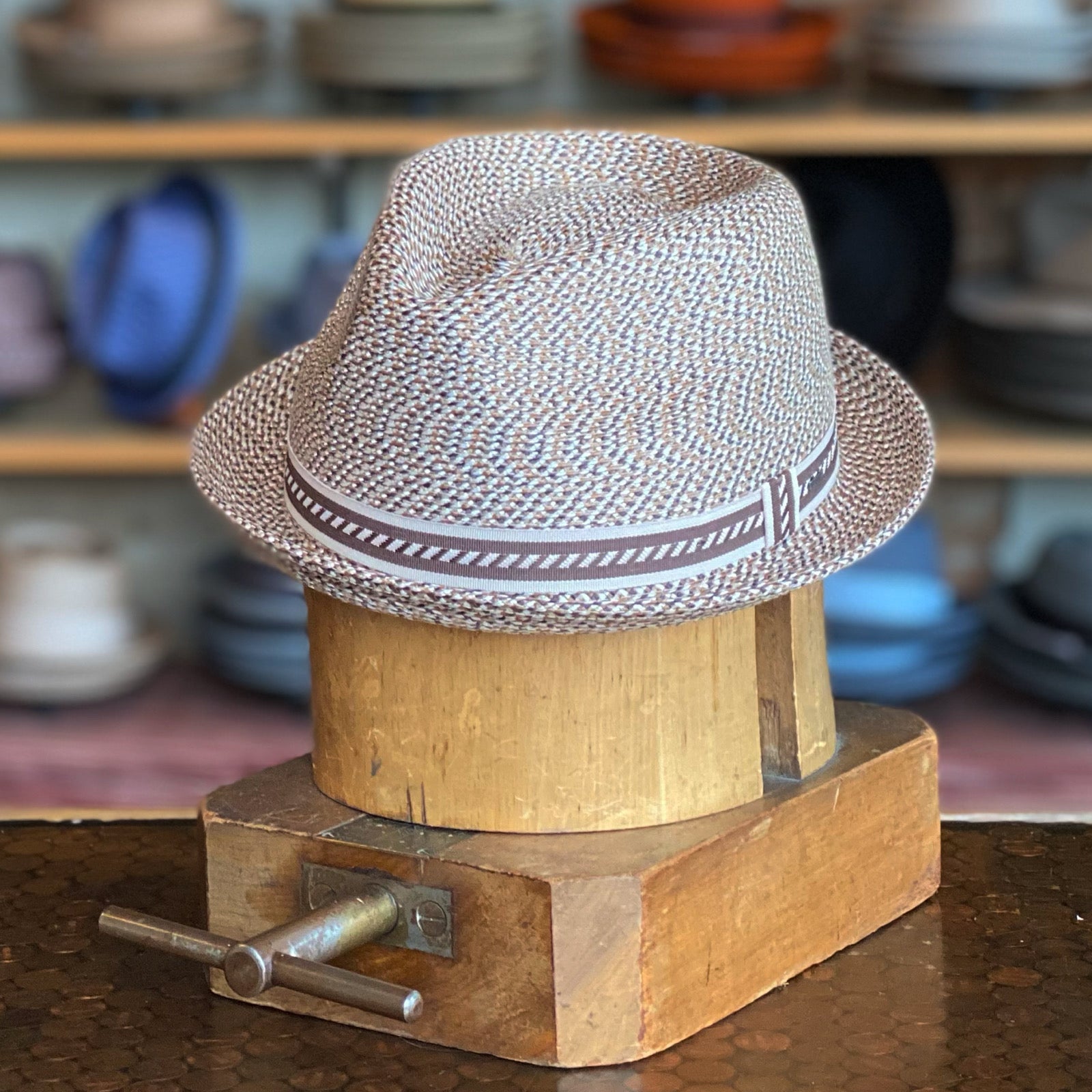 Mannes hat displayed on a wooden hat block, featuring a light taupe woven material with a matching patterned band, styled as a classic fedora hat.