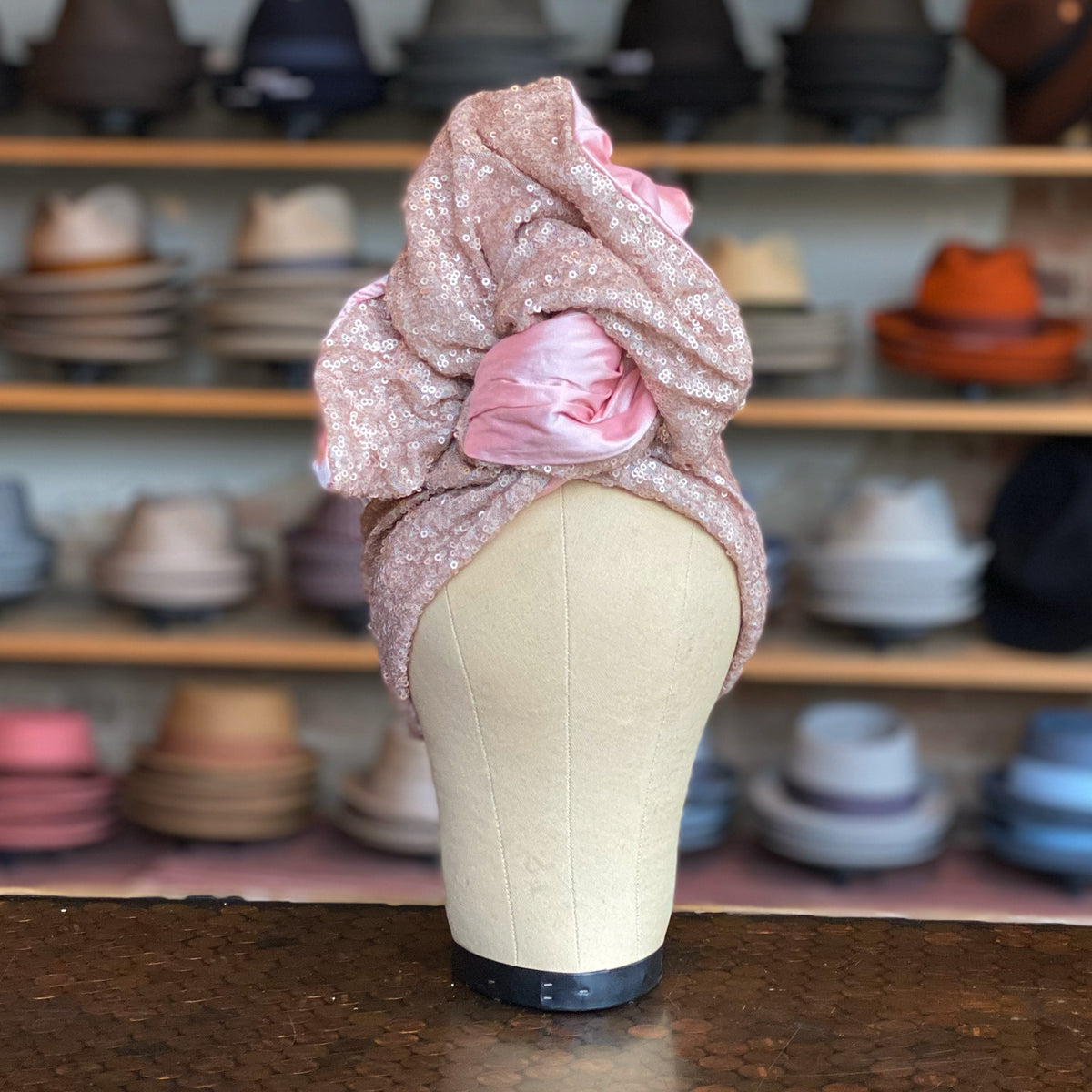 Sequin Twisturban turban-style hat in pink fabric with sparkling sequins and a twisted knot detail, displayed on a mannequin against a background of various hats.