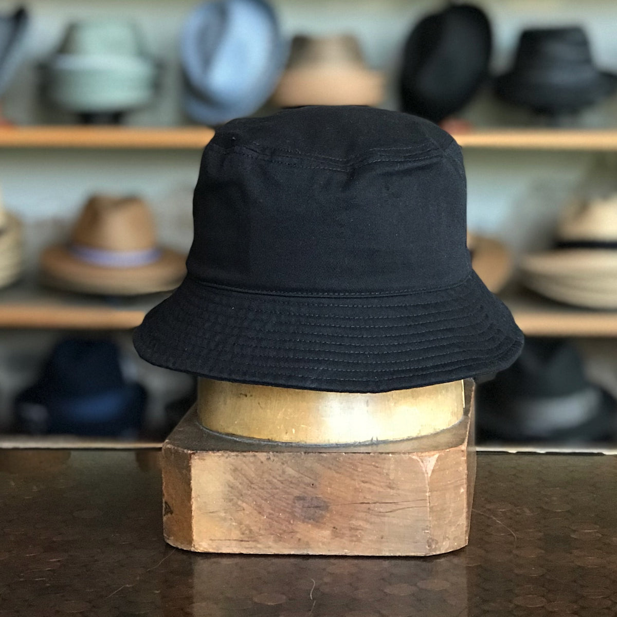 Black Washed Bucket Hat displayed on a wooden block with various hats in the background, showcasing a classic brim on fifth style accessory.