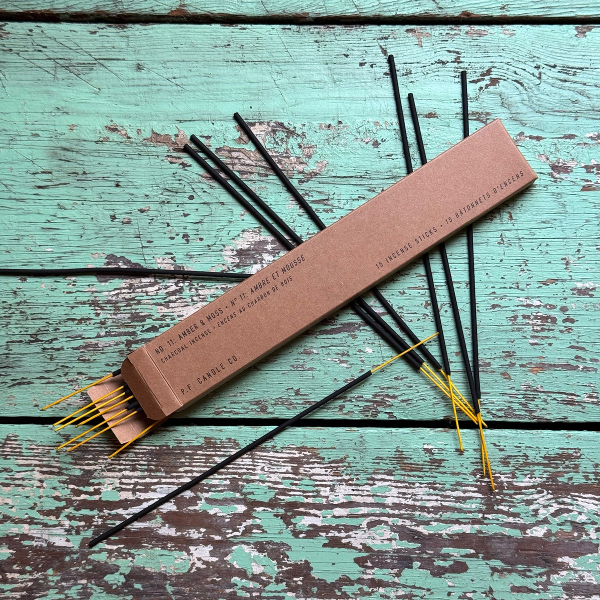 Amber & Moss Incense sticks arranged on a weathered turquoise wooden surface with a simple brown packaging box, highlighting a natural and rustic aesthetic.