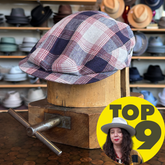 Americana Vintage Flat Cap in a plaid pattern with shades of red, white, and navy, displayed on a wooden hat block in a hat shop showcasing various hats in the background.