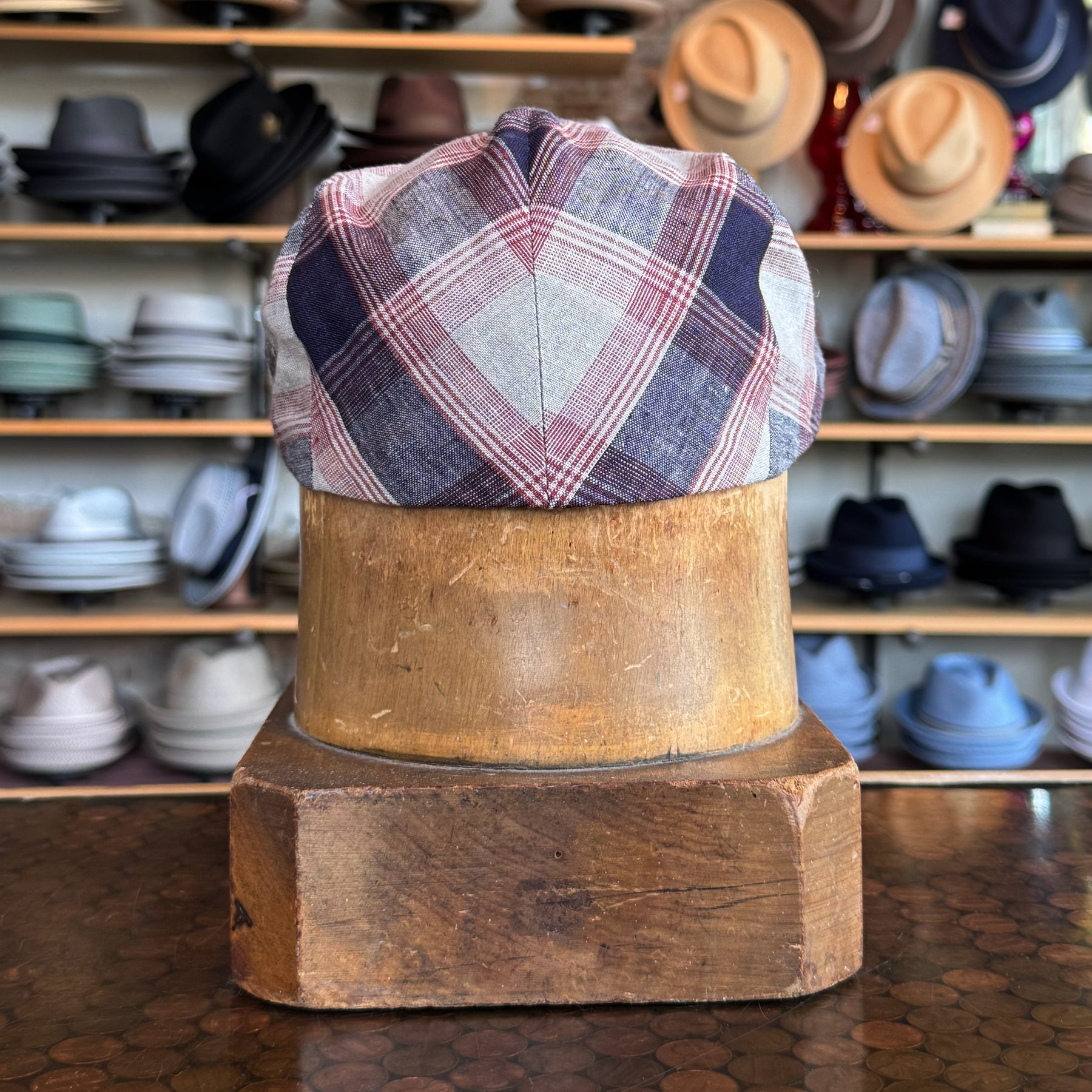 Back view of Americana Vintage Flat Cap in a plaid pattern with navy, red, and gray tones displayed on a wooden hat block in a store with various hats in the background.