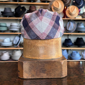 Back view of Americana Vintage Flat Cap in a plaid pattern with navy, red, and gray tones displayed on a wooden hat block in a store with various hats in the background.