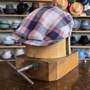 Americana Vintage Flat Cap in a red, white, and blue plaid pattern displayed on a wooden hat block with various hats blurred in the background.