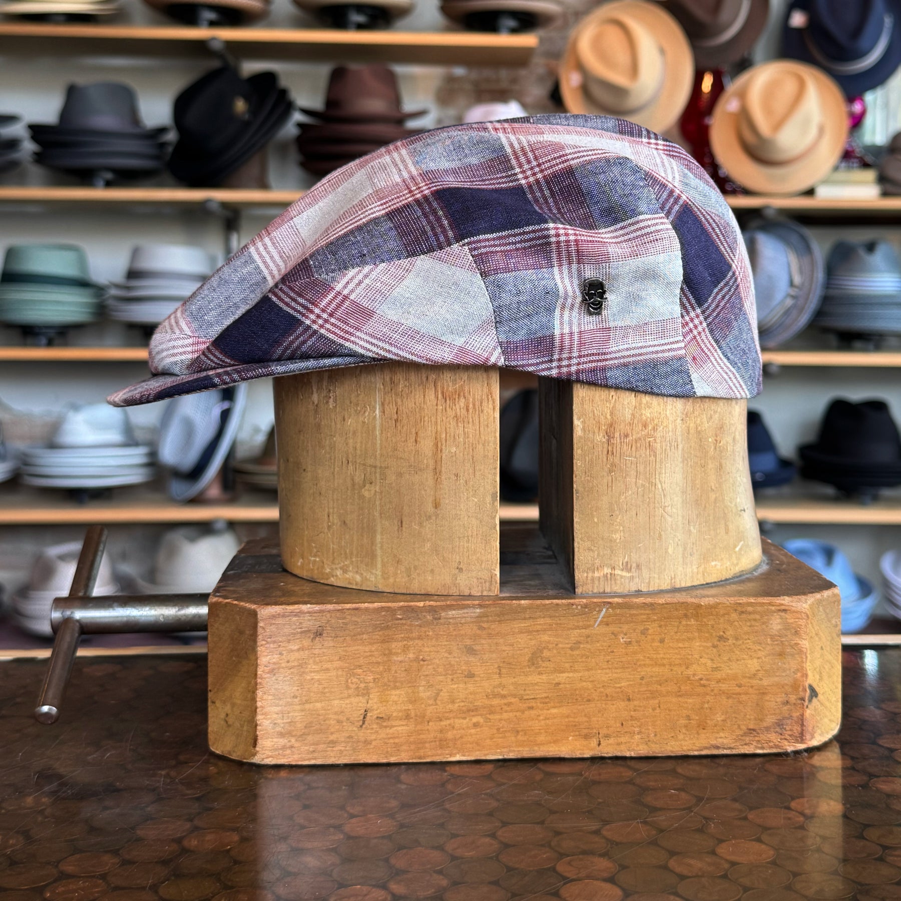 Americana Vintage Flat Cap in a red, blue, and white plaid pattern displayed on a wooden hat block with shelves of various hats in the background.