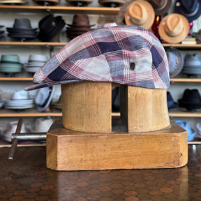 Americana Vintage Flat Cap in a red, blue, and white plaid pattern displayed on a wooden hat block with shelves of various hats in the background.
