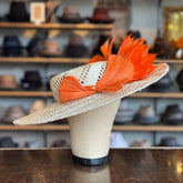 Straw Apple Blossom Boater hat with a wide brim and decorative burnt orange bow and feathers, displayed on a beige hat stand in a hat store.