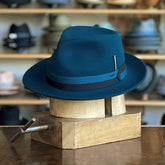 Side of Appley wool  Fedora in a sea moss color with a teal and black band, displayed on a wooden hat block in a boutique setting featuring various hats in the background.