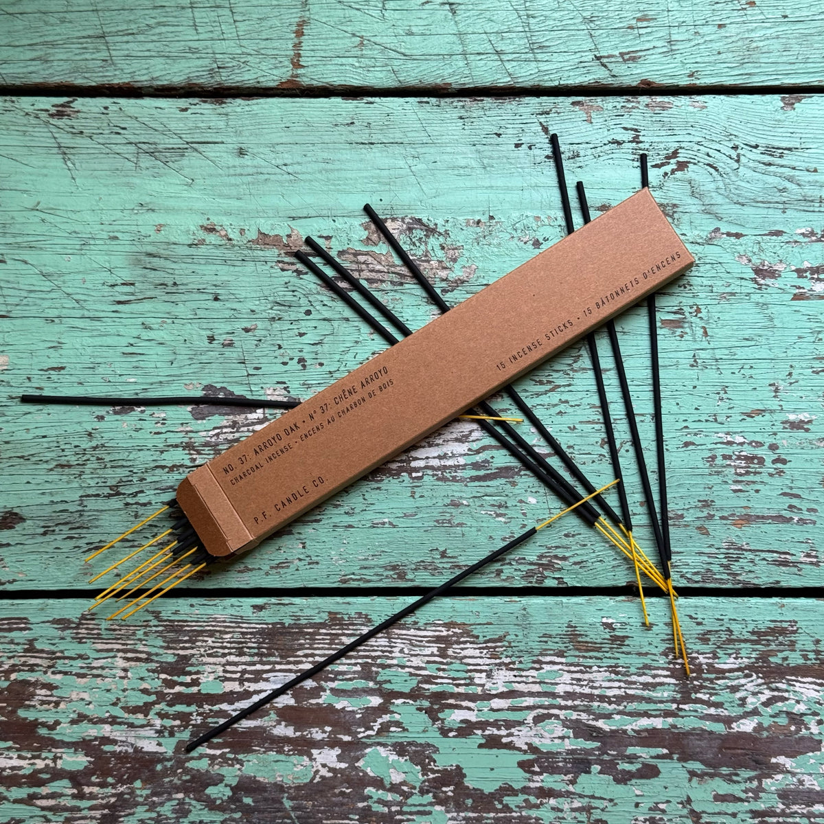 Arroyo Oak Incense sticks with black stems and yellow tips arranged on a distressed turquoise wooden surface next to a rectangular brown cardboard box.