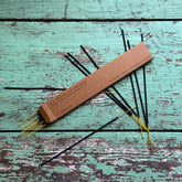 Arroyo Oak Incense sticks with black stems and yellow tips arranged on a distressed turquoise wooden surface next to a rectangular brown cardboard box.
