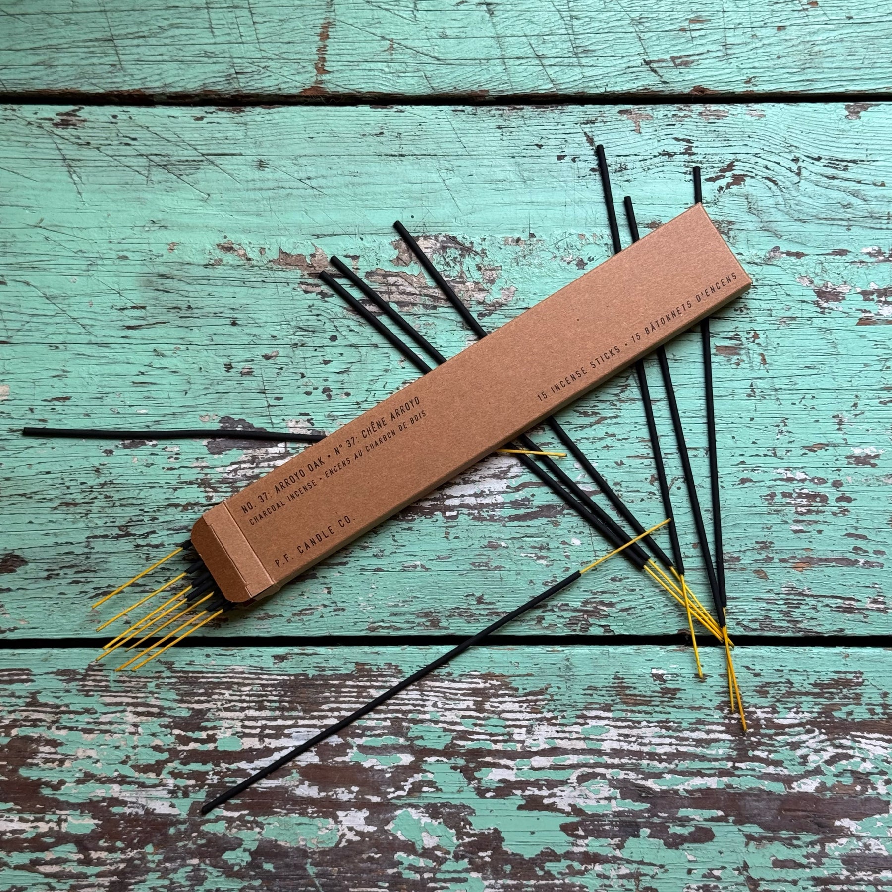 Arroyo Oak Incense sticks with black stems and yellow tips arranged on a distressed turquoise wooden surface next to a rectangular brown cardboard box.