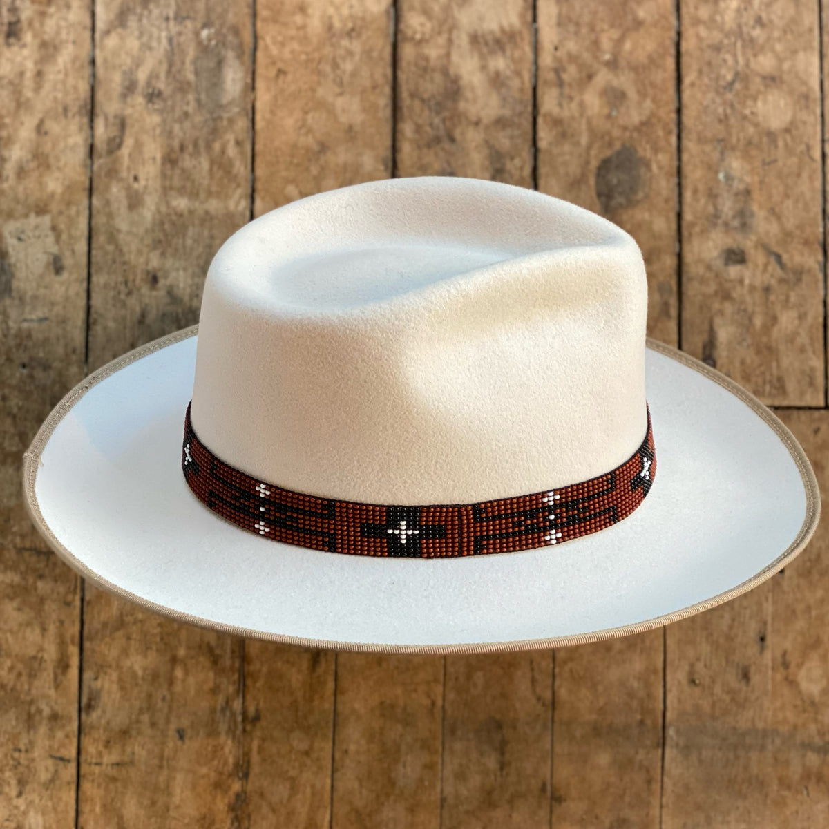 White felt hat featuring a decorative Beaded Hat Band with a geometric pattern in brown, black, and white, styled on a wooden surface background.