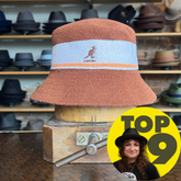 Bermuda Stripe Bucket Hat in a textured brown material with a light blue and orange stripe band featuring a small kangaroo logo, displayed on a wooden stand in a hat shop.