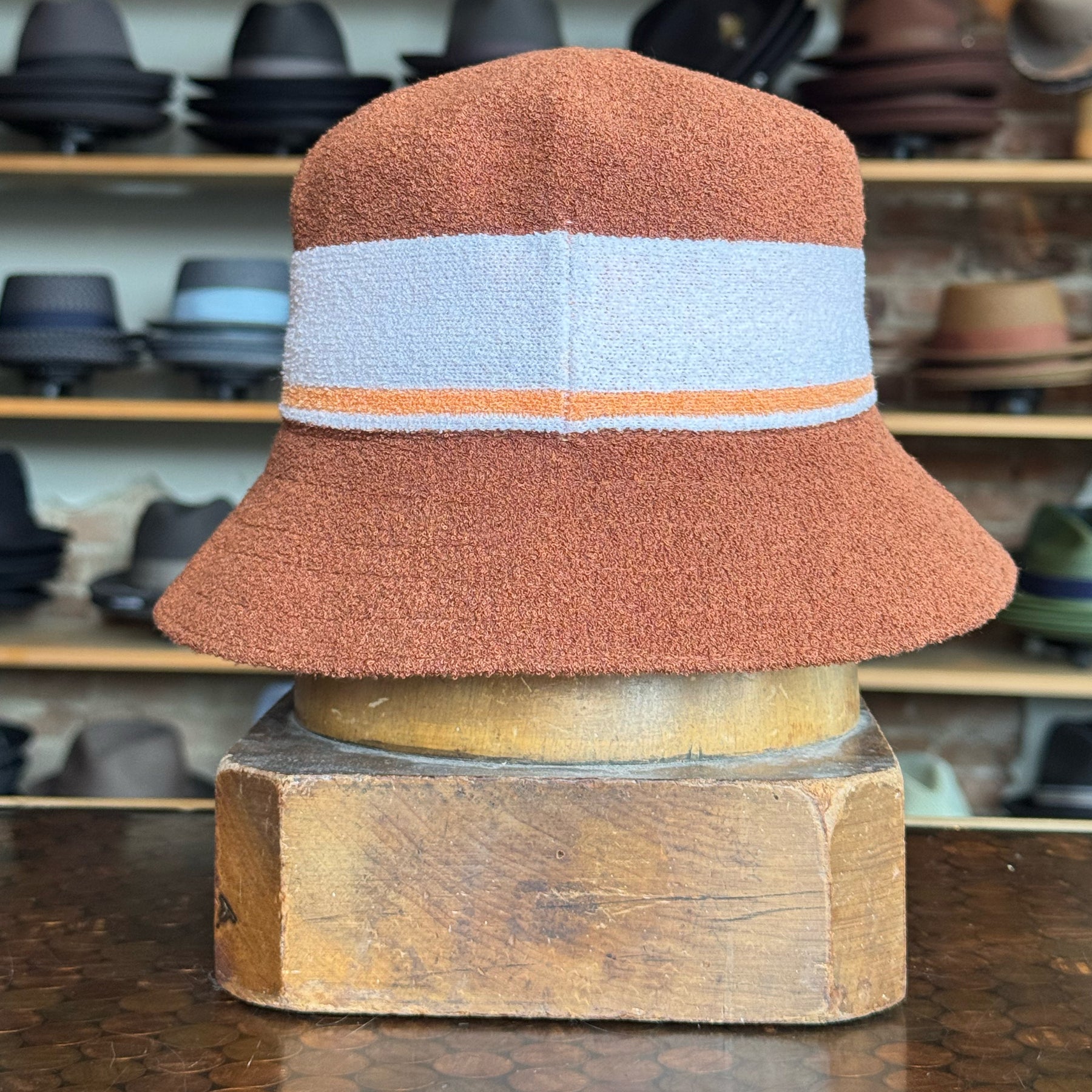 Bermuda Stripe Bucket Hat in a textured mahogany color with a wide light blue and thin orange stripe around the crown, displayed on a wooden stand with other hats blurred in the background.
