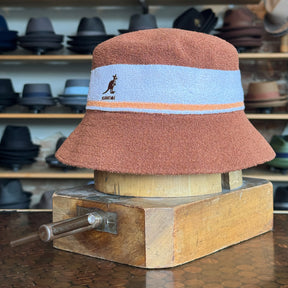 Bermuda Stripe Bucket Hat in a soft textured fabric with mahogany color and light blue and orange stripes, displayed on a wooden stand in a hat shop.