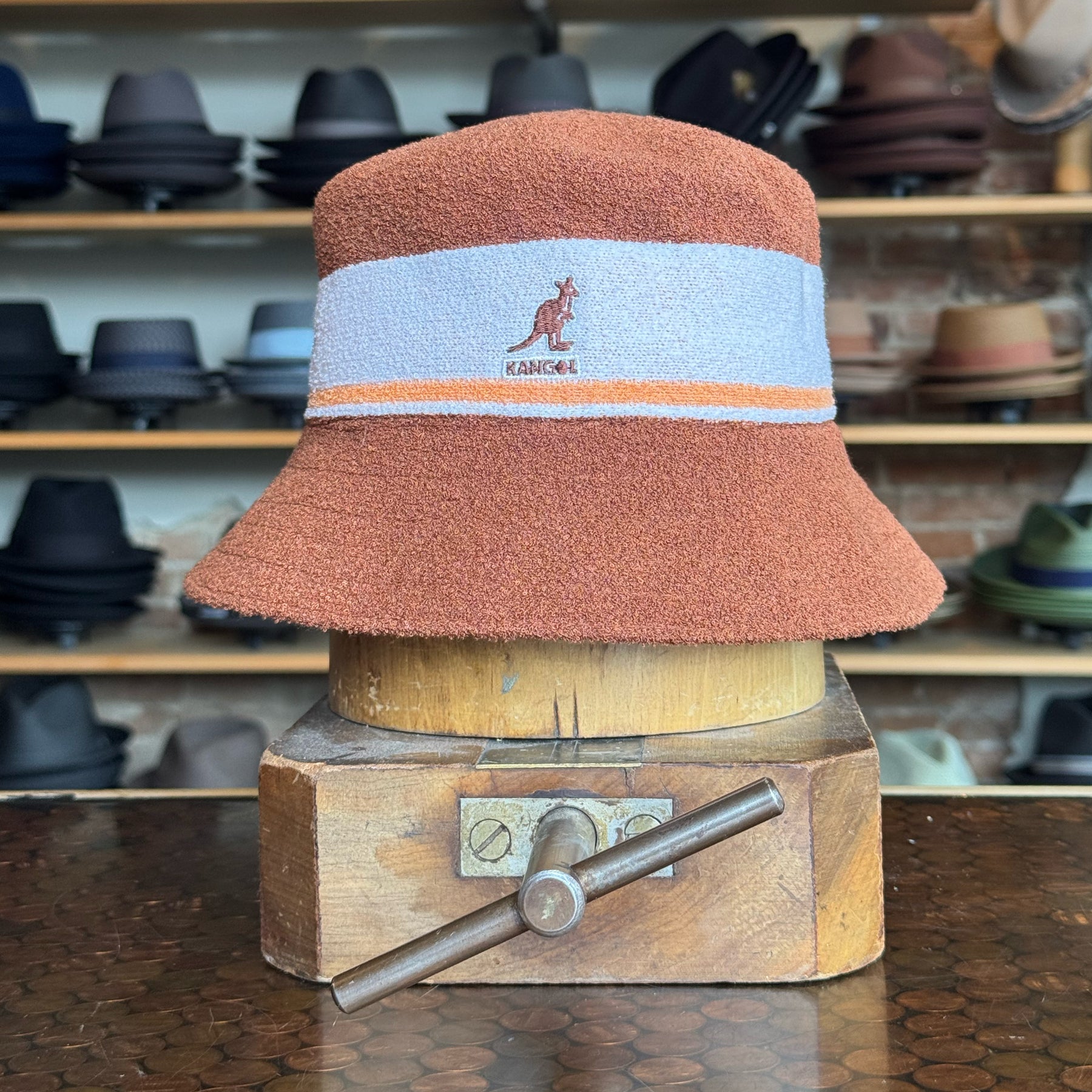 Bermuda Stripe Bucket Hat in warm mahogany and light blue with orange stripe detail, displayed on a wooden hat block in a shop filled with various brim hats.