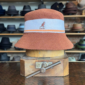 Bermuda Stripe Bucket Hat in warm mahogany and light blue with orange stripe detail, displayed on a wooden hat block in a shop filled with various brim hats.