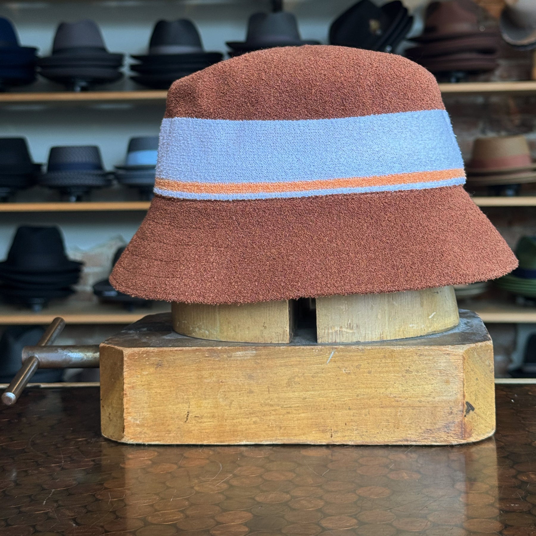 Bermuda Stripe Bucket Hat in a textured mahogany brown fabric with a wide blue and orange stripe detail, displayed on a wooden hat block surrounded by various brim hats.