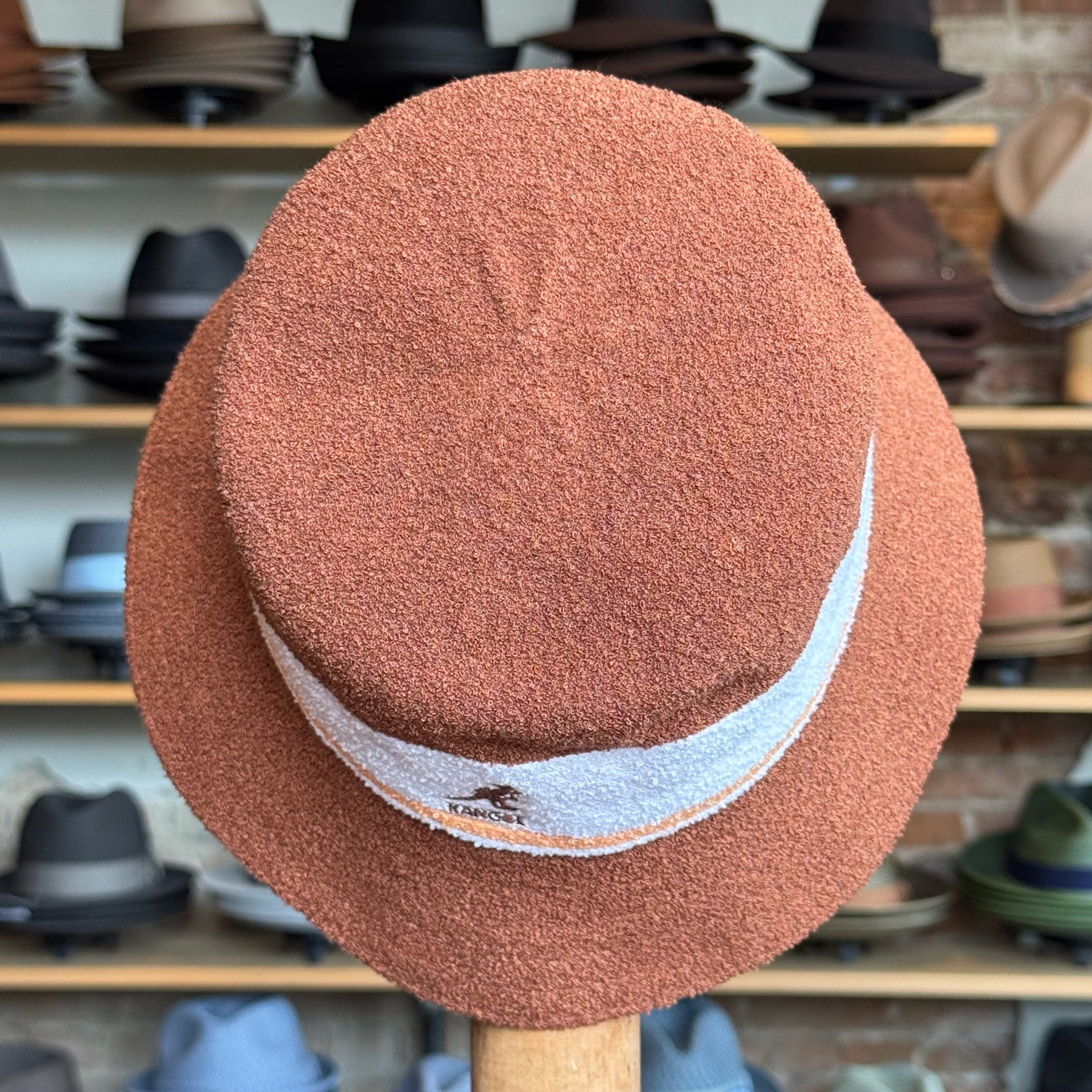 Bermuda Stripe Bucket Hat in warm mahogany with a textured fabric and a contrasting white stripe accented with subtle branding, displayed in a hat shop setting with various hats in the background.