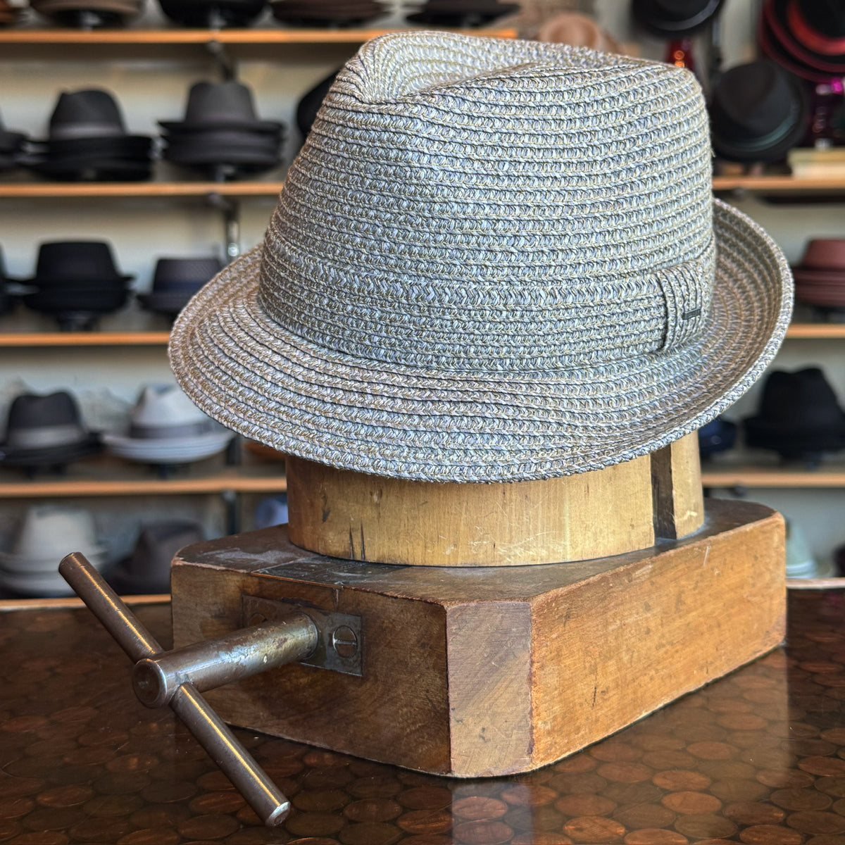 Billy flat cap made of woven grey material displayed on a wooden hat block in a shop with various hats in the background