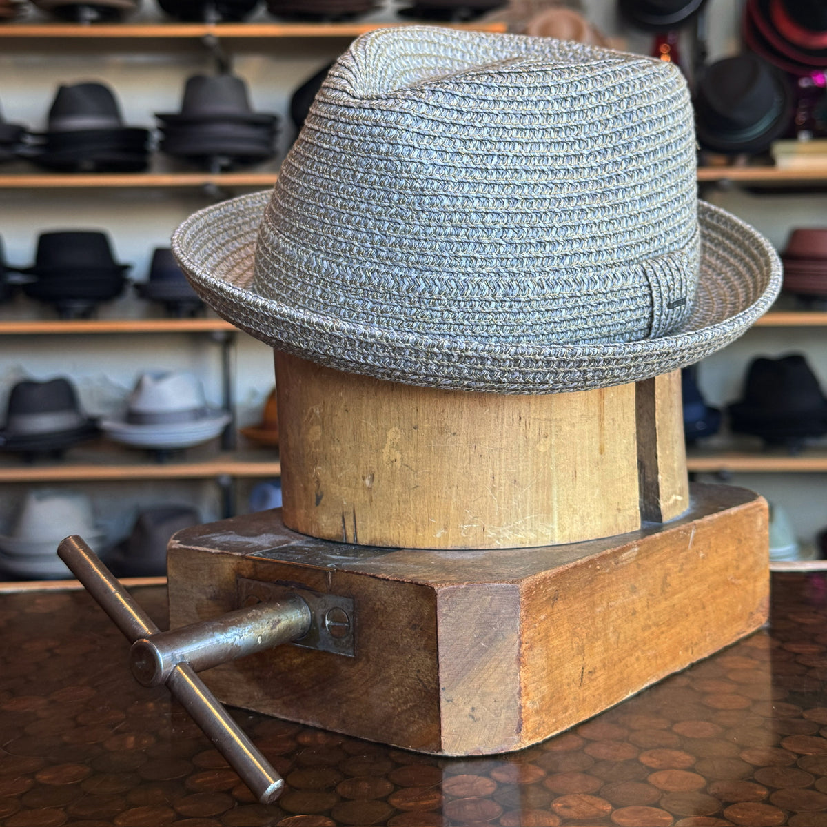 Billy hat made of woven grey material displayed on a wooden hat block with various hats blurred in the background.