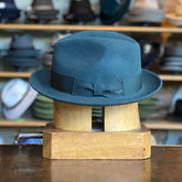 Side of Blixen flat cap in a deep sea moss green tone with a matching grosgrain band, displayed on a wooden hat block against a backdrop of various hats in a shop setting.