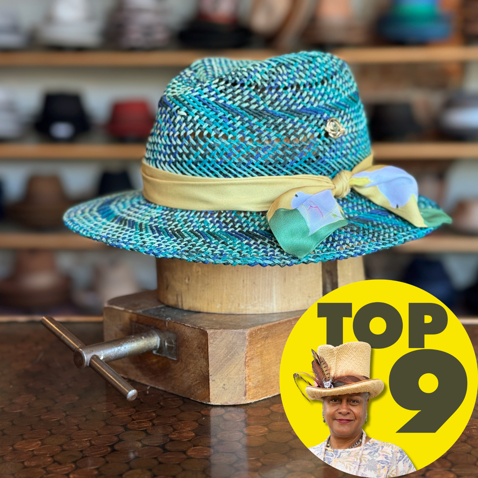 Bunny Hop Fedora made of woven blue, turquoise, and gray straw with a beige ribbon and decorative pastel scarf tied on the side, displayed on a wooden stand.
