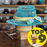 Bunny Hop Fedora made of woven blue, turquoise, and gray straw with a beige ribbon and decorative pastel scarf tied on the side, displayed on a wooden stand.