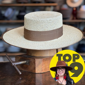 Canero Bolero straw hat with a wide brim and brown band, displayed on a wooden hat block in a rustic setting with shelves of hats blurred in the background.