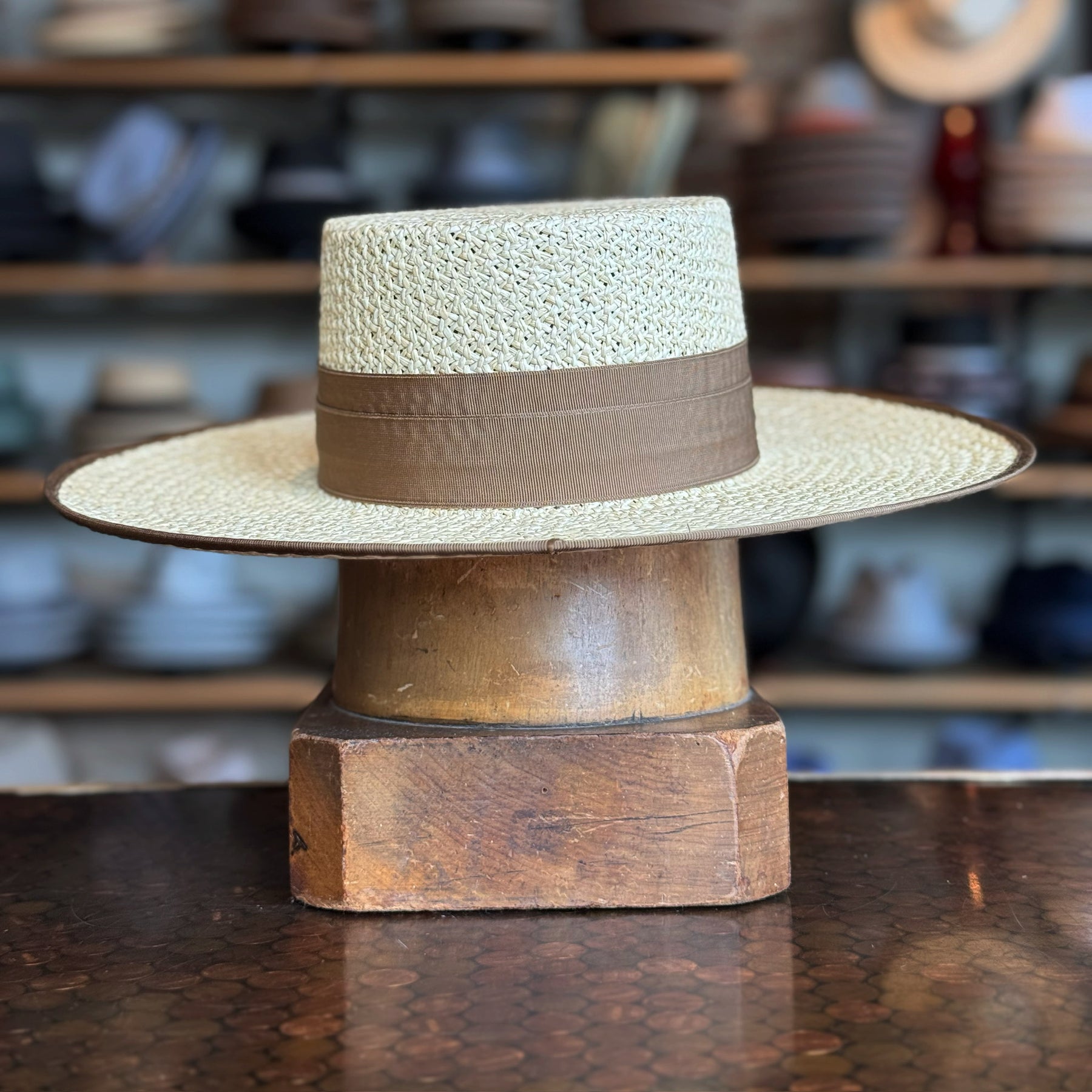 Canero Bolero straw hat with wide brim and brown band displayed on a wooden hat stand, set against a blurred background of various hats and accessories.